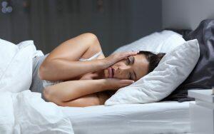 A woman experiencing morning jaw pain rests her hand on her head while lying in bed.