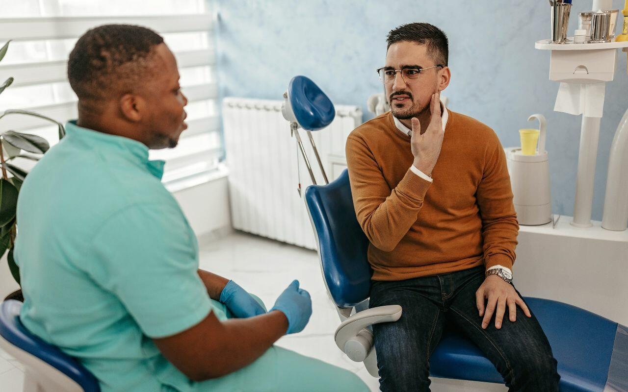 A man sitting in a dentist's chair talking to another man.